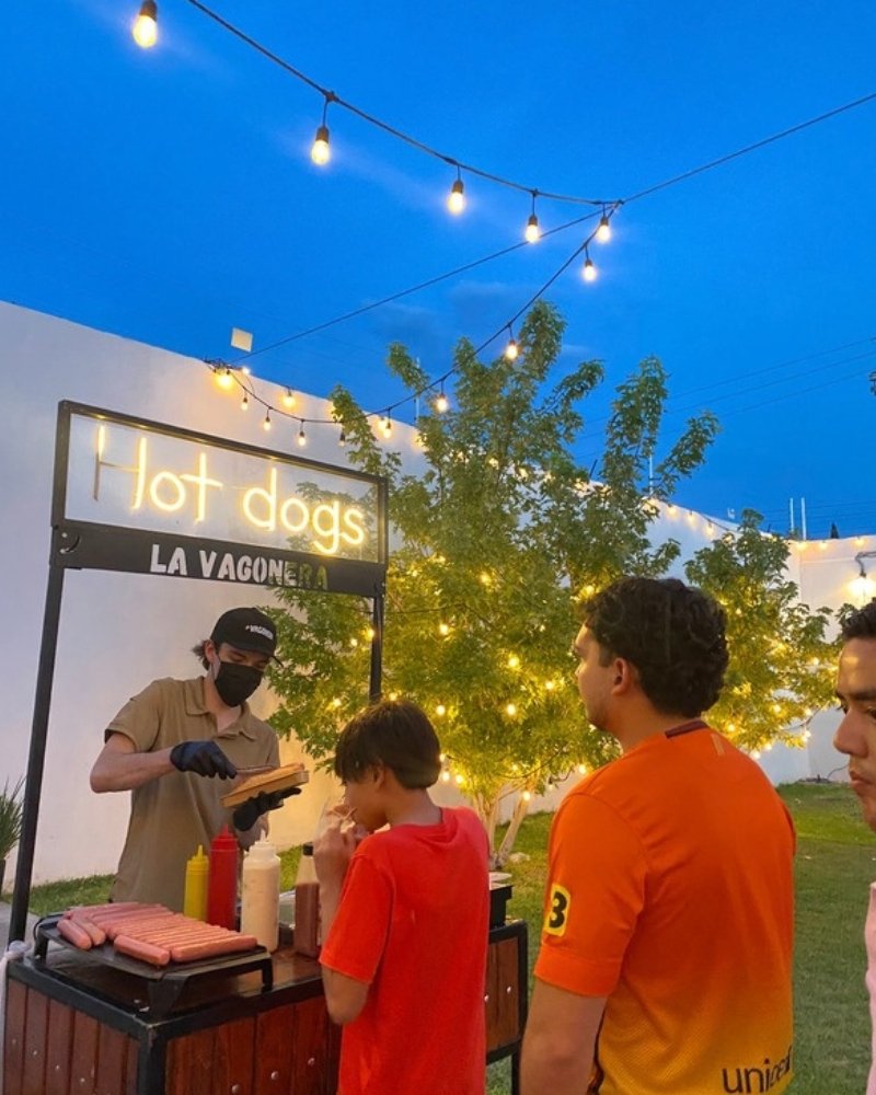 Carrito de hot dogs de La Vagonera al atardecer en un jardín con luces